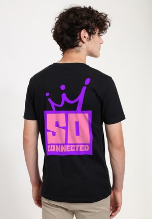 Ung mand med krøllet hår vender sidelæns iført sort t-shirt med stor pink og lilla tekst "SO CONNECTED" og kronegrafik på ryggen.