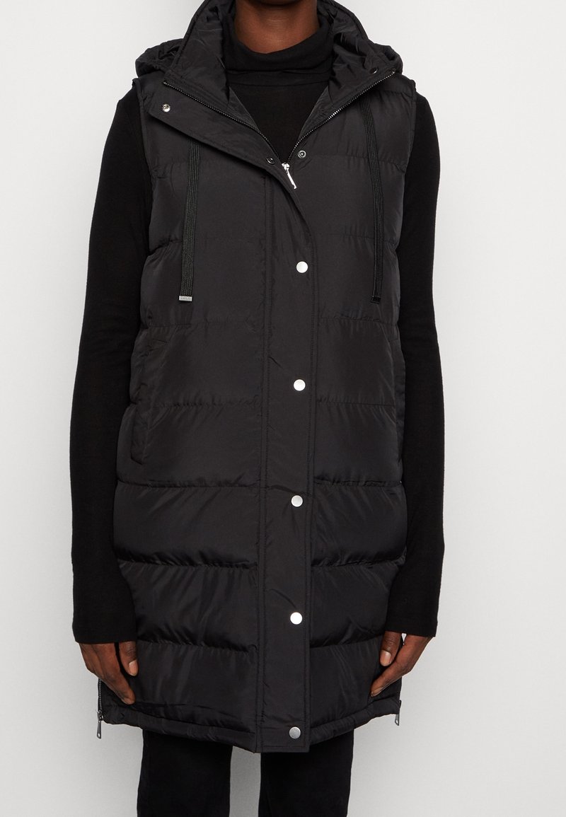 Personne portant un gilet matelassé noir sans manches avec des boutons-pression et une capuche par-dessus un pull noir à col roulé et manches longues.