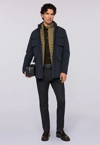 Tmavě modrá vnější bunda s několika kapsami vpředu, přes světlohnědou prošívanou vestu, v kombinaci s tmavými džínami a černými botami. Drží černou tašku.