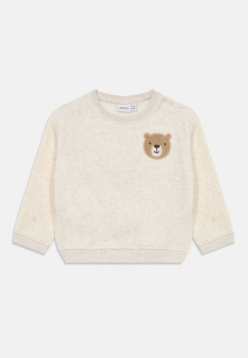 Felpa beige per bambino con maniche lunghe, polsini e orlo a coste, bottoni a pressione sulla spalla e una piccola toppa a forma di faccia di orsacchiotto sul petto.