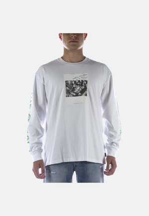 Homme portant une chemise blanche à manches longues avec un motif noir et blanc sur la poitrine et un texte vert imprimé sur les manches, porté avec un jean clair déchiré.