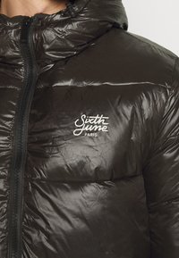 Doudoune en nylon noir avec une texture brillante, dotée d'un col montant, d'une fermeture éclair frontale, et du logo blanc "Sixth June Paris" sur la poitrine.