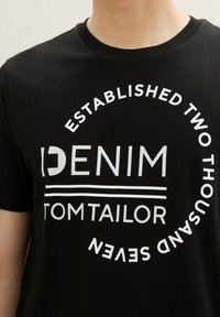 Čierne bavlnené tričko s okrúhlym bielym potlačou s textom "DENIM" a "TOM TAILOR", a detailom "ZALOŽENÉ DVA TISÍC SEDMÝ".