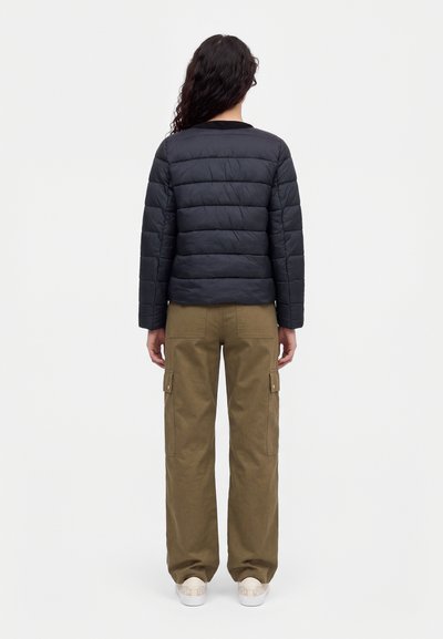 Une personne se tient de dos et porte une veste noire matelassée, un pantalon cargo olive et des baskets blanches sur un fond uni.