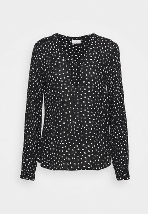 Blouse noire à manches longues avec des pois blancs irréguliers et un décolleté en V sur un fond uni.