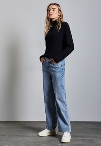 Suéter de cuello alto acanalado en azul marino combinado con jeans de pierna ancha en azul claro y zapatillas blancas, ubicado frente a un fondo gris neutro.