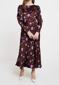 Brun blommig klänning med långa puffärmar, prydd med lila blommor och en flödande design. Bärs med vita klackar mot en enkel bakgrund.