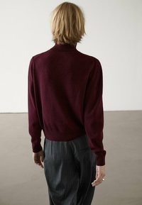 Person mit kurzen blonden Haaren, die von hinten zu sehen ist, trägt einen burgunderfarbenen Rollkragenpullover und dunkle gestreifte Hosen in einem minimalistischen Innenraum.