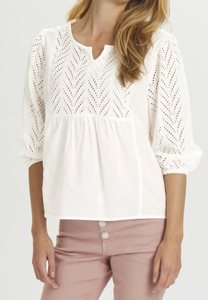 Vrouw die een witte oogjeskant blouse draagt met driekwart pofmouwen en mauve broek met decoratieve knopen.