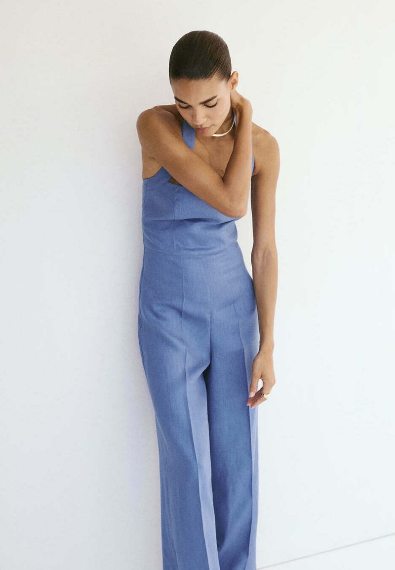 Femme avec un chignon sleek porte une combinaison bleu sans manches, debout contre un mur blanc avec une main touchant son cou.