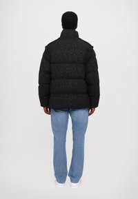 Black puffer jacket with a textured pattern, high collar, and a drop-shoulder design, paired with light blue jeans and white sneakers.