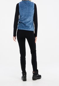Person som bär en blå fleeceväst, svart långärmad topp, svarta figurnära byxor och svarta vandringskängor, stående mot en vit bakgrund.
