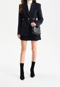 Blazer marine avec une ceinture, associé à un pull col roulé gris et une jupe marine. Des bottines noires en suède et un petit sac à main noir complètent le look.