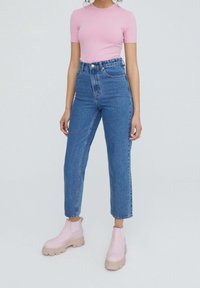 Top corto a maniche corte di un rosa chiaro a coste abbinato a jeans in denim blu a vita alta e stivaletti rosa con tacco chunky. Sfondo neutro.