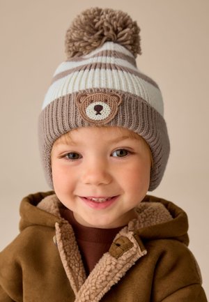 Niño sonriente con un abrigo marrón forrado de forro polar y un gorro de punto a rayas con pompón y un parche de oso.