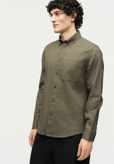 Homme portant une chemise boutonnée olive à manches longues avec une poche poitrine, debout de profil sur un fond clair uni.