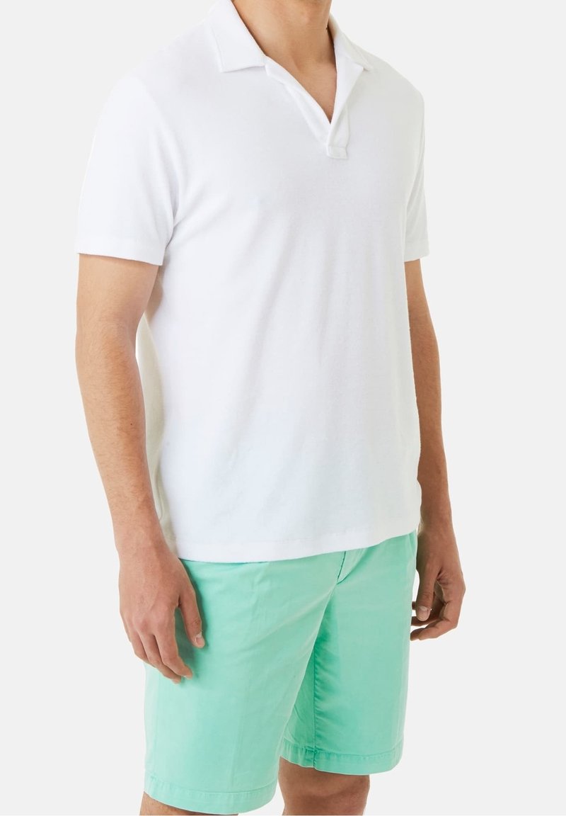 Homme portant un polo blanc à manches courtes et un short vert menthe, debout devant un fond clair uni.