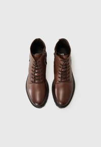 Chaussures en cuir marron avec un bout rond, dotées de lacets et de fermetures éclair, disposées sur un fond clair.