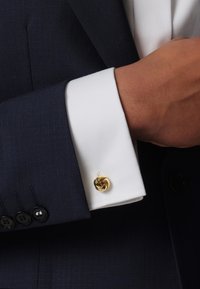 Navy blue suit jacket with a white cuff featuring a gold spiral cufflink. Jacket has a textured fabric; cuff is smooth and crisp.
