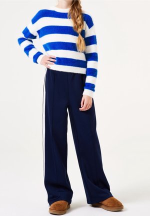 Personne portant un pull en tricot duveteux à rayures bleues et blanches, un pantalon large bleu marine avec des bandes blanches sur les côtés, et des chaussures marron sans lacets, debout avec la main sur la hanche.