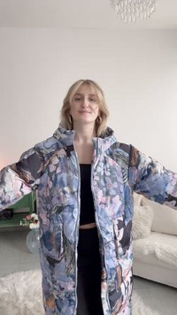 Junge Frau trägt eine gepolsterte Jacke mit pastellfarbenem Blumenmuster, steht mit ausgebreiteten Armen in einem gut beleuchteten Wohnzimmer.