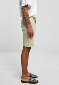 Shorts beige de mezcla de algodón, con corte recto. Llevados con sandalias negras tipo slide. Tatuajes visibles en el antebrazo.