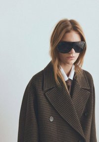 Abrigo marrón a cuadros de doble botonadura con grandes solapas con muesca, combinado con camisa blanca y corbata marrón. Gafas de sol negras extragrandes completan el conjunto.