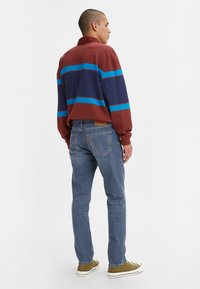 Camisa polo de manga larga colorida con rayas en burdeos, azul y marino, combinada con jeans azules y zapatillas de deporte en verde oliva, vista desde atrás.
