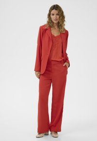 Femme aux cheveux bouclés portant un débardeur en dentelle rouge, un blazer rouge assorti et un pantalon large, se tenant avec une main dans la poche.