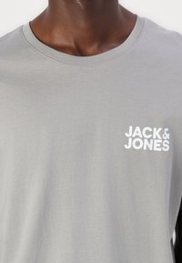 Graues Kurzarm-T-Shirt aus weichem Stoff, mit rundem Halsausschnitt und einem weißen Logo auf der linken Brustseite.