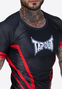 Rash guard negro de manga corta con un logo blanco de "TAPOUT". Los acentos rojos y el diseño salpicado crean un aspecto dinámico. Material suave y ajustado.
