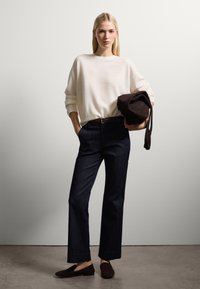 Suéter de punto crema, jeans rectos de mezclilla azul oscuro, zapatos de ante marrón. Sosteniendo un bolso de ante marrón con correas delgadas. Diseño simple y moderno.