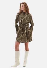 Robe de couleur vert olive avec un motif paisley, à manches longues, taille cintrée et col haut, associée à des bottes blanches jusqu'aux genoux.