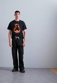 Black t-shirt with a bear graphic in orange flames, paired with black cargo pants featuring white side stripes and black shoes.