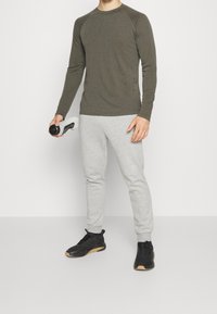 Olivgrünes Langarmshirt, graue Jogginghose und schwarze Sportschuhe. Hält eine weiße Wasserflasche mit schwarzen Akzenten.