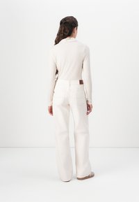 Kremfarget, ribbestrikket langermet topp i kombinasjon med høyhalsede, vide hvite jeans. Outfiten har en subtil brun lærdetalj ved midjen.