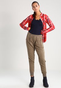 Chemise en flanelle à carreaux rouges et noirs sur un débardeur côtelé bleu marine, associée à un pantalon tapered kaki et des bottines noires, design minimaliste.