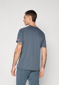 Mann mit kurzem, dunklem Haar und Tattoos, der ein graues, kurzärmeliges Hemd und passende Hosen trägt, mit dem Rücken zu einer schlichten Hintergrund.