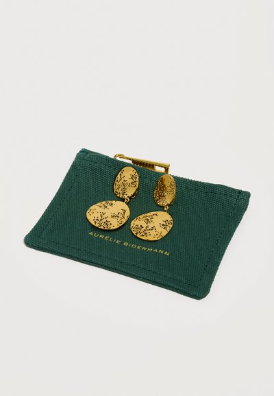 Χρυσά οβάλ σκουλαρίκια με σταγόνα και λεπτές φυτικές χαραγές που ακουμπούν σε τσαντάκι από σκούρο πράσινο ύφασμα με χρυσή αγκράφα.