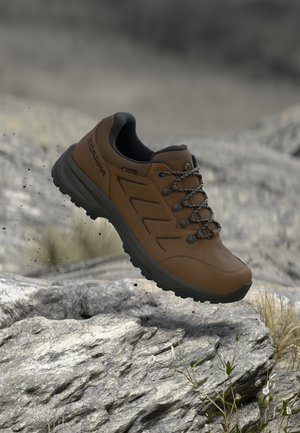 Hnědá treková bota s černou podrážkou a vzorovanými tkaničkami, která se vznáší nad kamenitou krajinou s řídkou trávou.