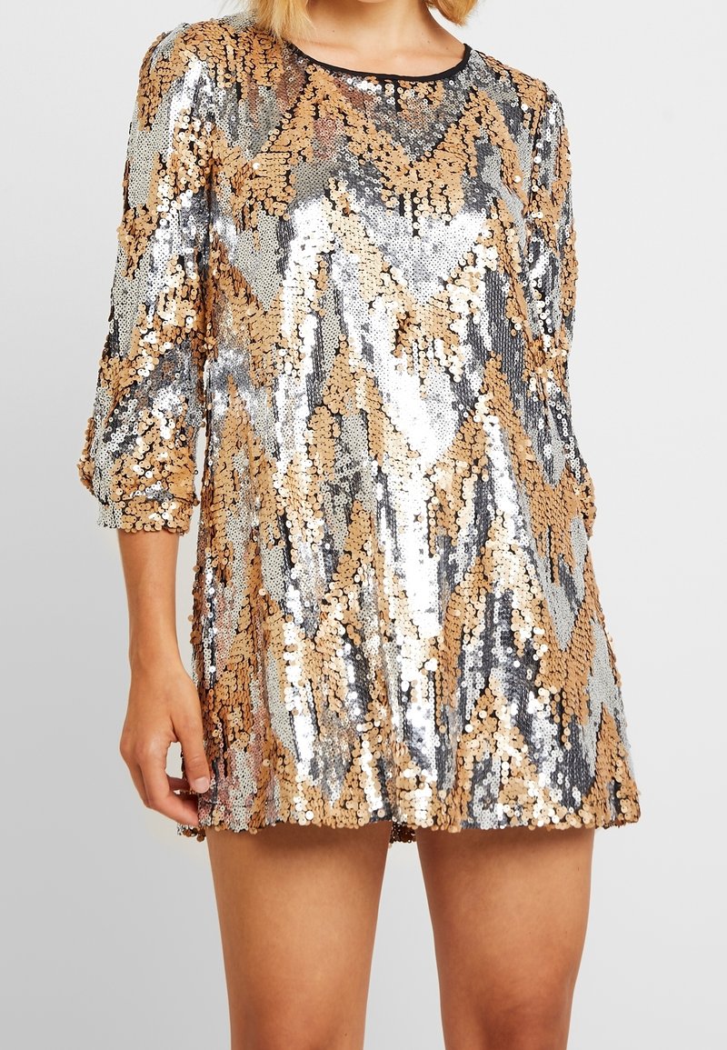 Femme portant une mini-robe brillante ornée de sequins chevrons dorés et argentés, avec des manches trois-quarts et un col rond.