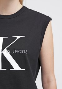 Zwarte mouwloze top met grote witte "K" en gedeeltelijke tekst "lein Jeans" zichtbaar op het bovenlichaam tegen een effen achtergrond.
