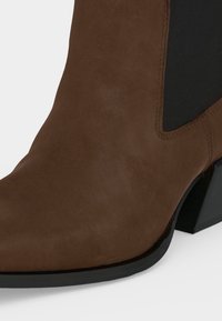 Bottine en suède marron avec panneau latéral élastique noir et petit talon bloc sur fond blanc.