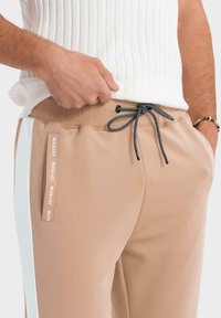 Beiga sportiga joggers med dragsko i midjan, sidofickor och vita accenter. Materialet har en slät textur med en åtsittande design.