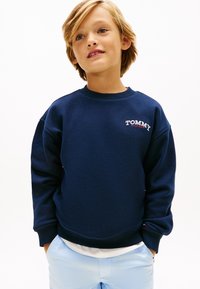 Μπλε σκούρο φούτερ με μακριά μανίκια, στρογγυλή λαιμόκοψη και λευκό λογότυπο "TOMMY HILFIGER" στο αριστερό στήθος. Συνδυασμένο με ανοιχτό μπλε παντελόνι.