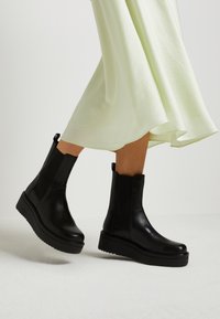 Botines de cuero negro con paneles elásticos laterales y suela de plataforma gruesa, combinados con un vestido vaporoso de color verde claro.