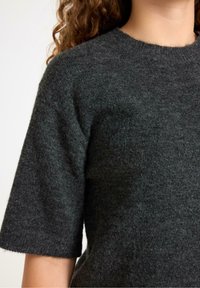 Ciemnoszara dzianinowa bluzka z krótkimi rękawami, okrągłym dekoltem i miękką, teksturowaną powierzchnią. Zbliżenie na górną część odzieży.