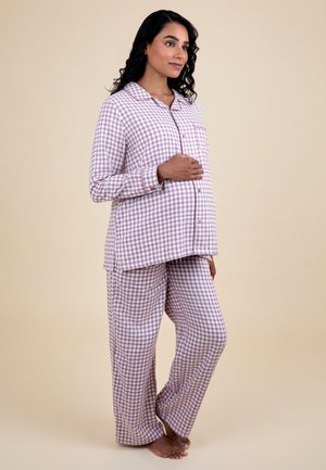 Tehotná žena stojaca naboso, oblečená v ružovo-biele kárované pyžamo s dlhým rukávom, s jednou rukou opierajúcou sa o brucho.