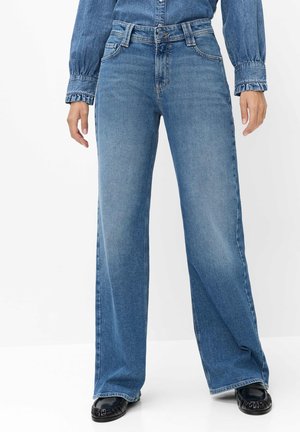 Kvinde iført blå jeans med vide ben, en denimskjorte med flæsekantede manchetter og sorte mokasin-sko, stående mod en hvid baggrund.