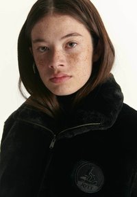 Chaqueta de pelaje sintético negro con cuello alto, cierre con cremallera y parche redondo de logo en el pecho. Textura suave y diseño moderno.